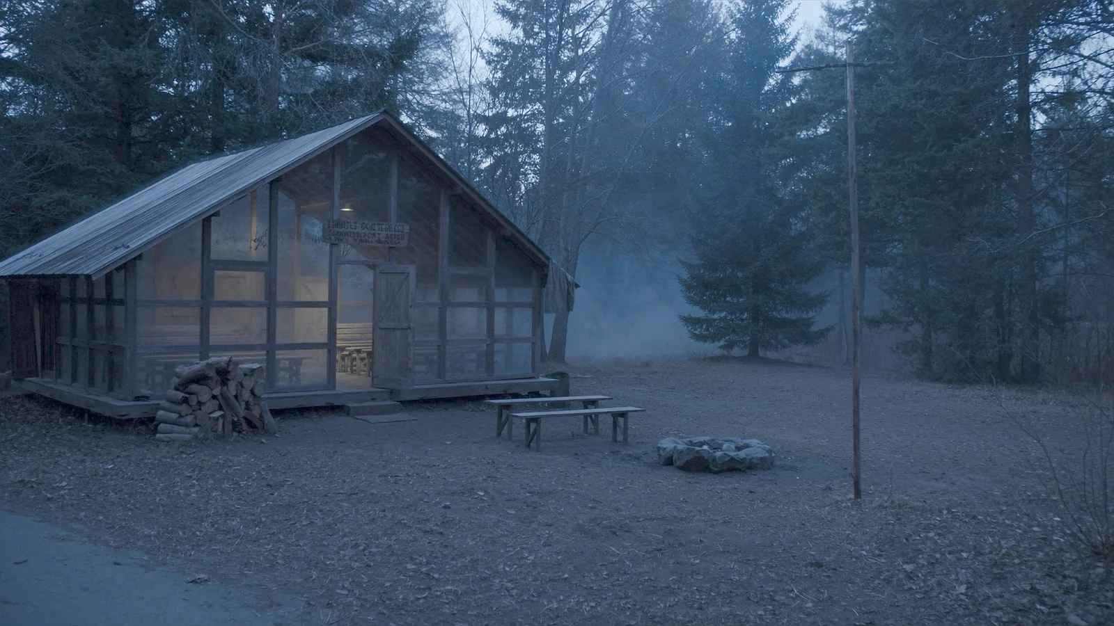 A weathered timber camp lodge and empty benches in a pine forest at dusk